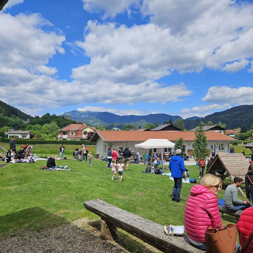 Eine Gemeinschaftsveranstaltung mit vielen Menschen auf einer Wiese. Es gibt Häuser, ein Zelt und Berge im Hintergrund. Die Leute sitzen, stehen und gehen herum.