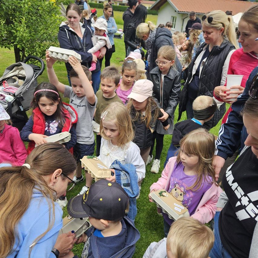 Eine Gruppe von Kindern und Erwachsenen versammelt sich auf einem Rasen, einige halten Eierkartons, während andere zuschauen. Im Hintergrund befinden sich Kinderwagen, Häuser und Bäume.