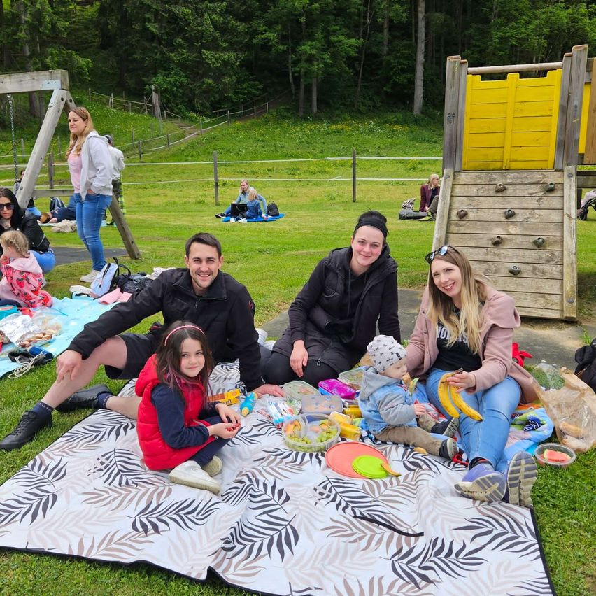 Eine vierköpfige Familie sitzt auf einer Picknickdecke in einem Park, umgeben von einer Spielstruktur und einer Schaukel. In der Nähe genießen andere Familien das Freie.