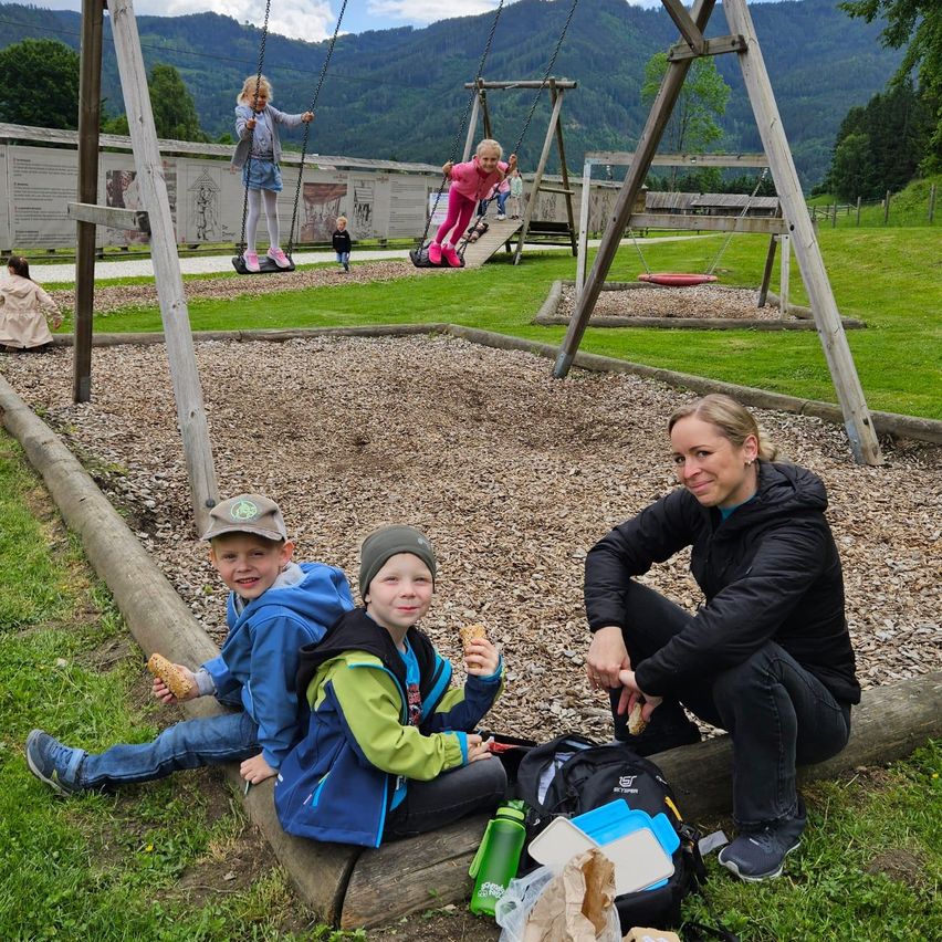 Eine Familie genießt ein Picknick auf einem Spielplatz mit Schaukeln und einer Rutsche, Berge im Hintergrund. Zwei Kinder sitzen und essen, eine Frau kniet neben ihnen.
