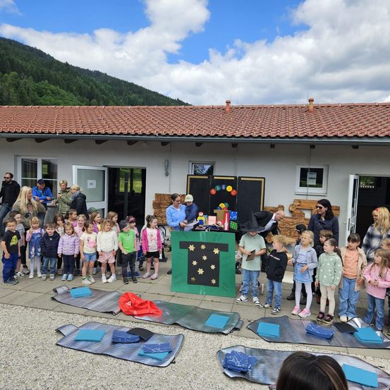Eine Gruppe von Kindern und Erwachsenen versammelt sich vor einem Gebäude mit einem Ziegeldach. Einige tragen Hüte und Schuhe. Ein grünes Banner mit einem gelben Sternenmuster ist ausgestellt.