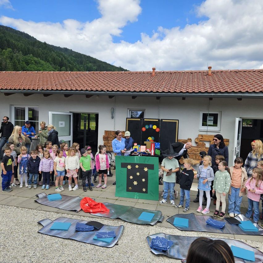 Eine Gruppe von Kindern und Erwachsenen versammelt sich vor einem Gebäude mit einem Ziegeldach. Einige tragen Hüte und Schuhe. Ein grünes Banner mit einem gelben Sternenmuster ist ausgestellt.