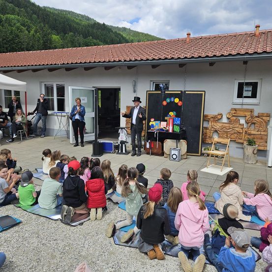 Ein Zauberer führt vor einer Gruppe von Kindern vor einem Gebäude mit rotem Dach draußen eine Vorstellung durch.