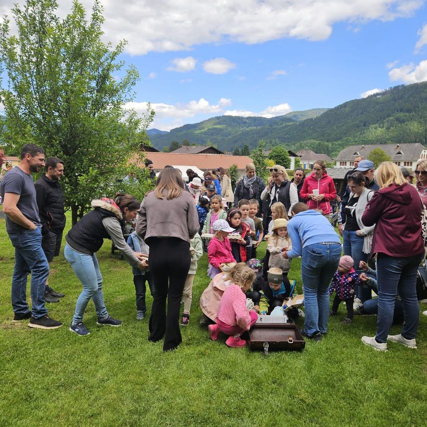Eine Gruppe von Menschen versammelt sich in einem Park. Einige Erwachsene beugen sich über einen Koffer, um den sich Kinder versammelt haben. Die Erwachsenen beobachten die Kinder. Der Himmel ist blau mit einigen Wolken.