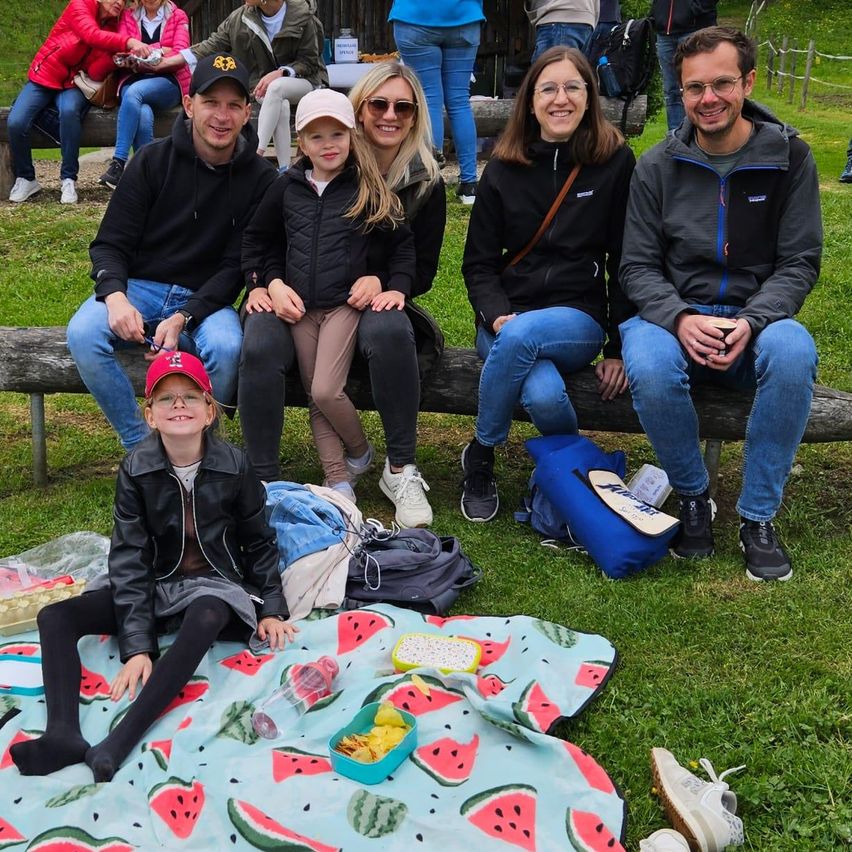 Eine Gruppe von Menschen sitzt auf einer Bank und einer Decke im Freien. Ein Kind sitzt mit Wassermelonen auf der Decke. Einige Leute stehen hinter ihnen. Einige Rucksäcke liegen auf dem Boden. Einige Essensartikel sind auf der Decke.
