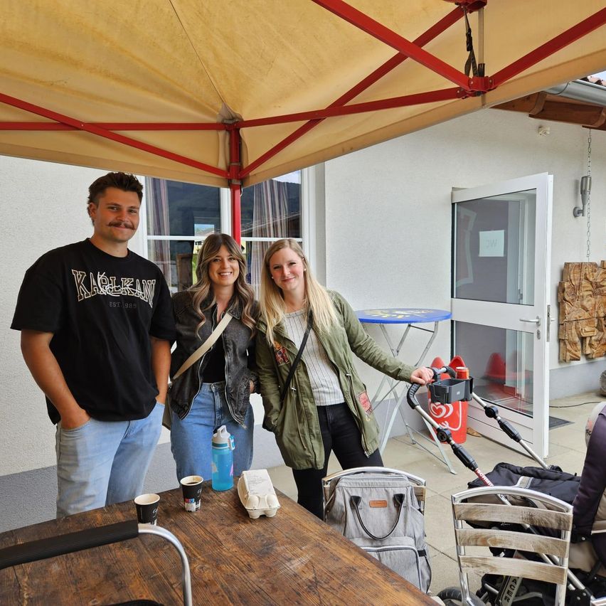 Drei Personen stehen unter einem Zelt mit weißem Baldachin, in der Nähe eines Tisches mit Tassen, Flaschen und einer Tasche. Eine Person hält einen Kinderwagen, und im Hintergrund befindet sich ein Stuhl und eine Tür.