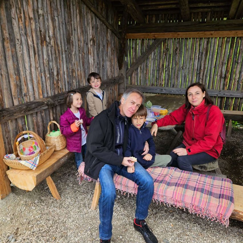 Eine Familie sitzt vor einem Holzschuppen und lächelt für ein Foto. Zwei Erwachsene sitzen auf einer Bank, während vier Kinder hinter ihnen stehen. Zwei Körbe stehen auf einem Tisch, und ein Picknicktisch mit Gegenständen befindet sich hinter den Erwachsenen.