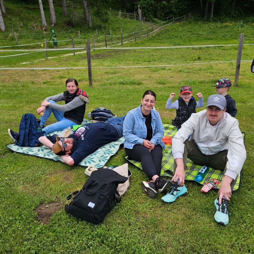 Eine Familie mit fünf Personen sitzt auf einem grünen Feld. Zwei Erwachsene sitzen auf der Decke, während zwei Kinder auf dem Gras sitzen. Ein Kind trägt einen Hut und das andere hält eine Flasche. Ein Rucksack liegt auf dem Boden. Das Gebiet ist von einem Zaun umgeben.