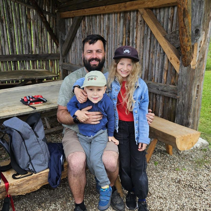 Eine dreiköpfige Familie posiert für ein Foto in einem Holzschuppen, ein Mann hält ein Kind und ein Mädchen steht neben ihm, mit Rucksäcken und einem Tisch im Hintergrund.