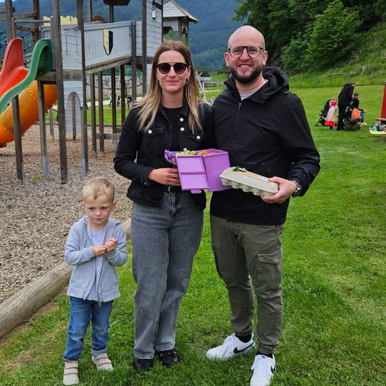 Eine Familie, bestehend aus einem Mann, einer Frau und einem kleinen Jungen, steht in einem Park. Sie halten eine lila Schachtel und Eierkartons. Hinter ihnen befindet sich ein Spielplatz mit Rutschen und einer burgartigen Struktur.