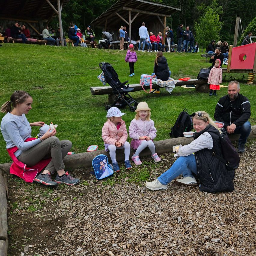 Eine Gruppe von Menschen, einschließlich Kindern, sitzt auf dem Boden in einem Park. In der Nähe gibt es Holzbänke und eine rote Spielstruktur. Das Gebiet ist von Gras und Bäumen umgeben.