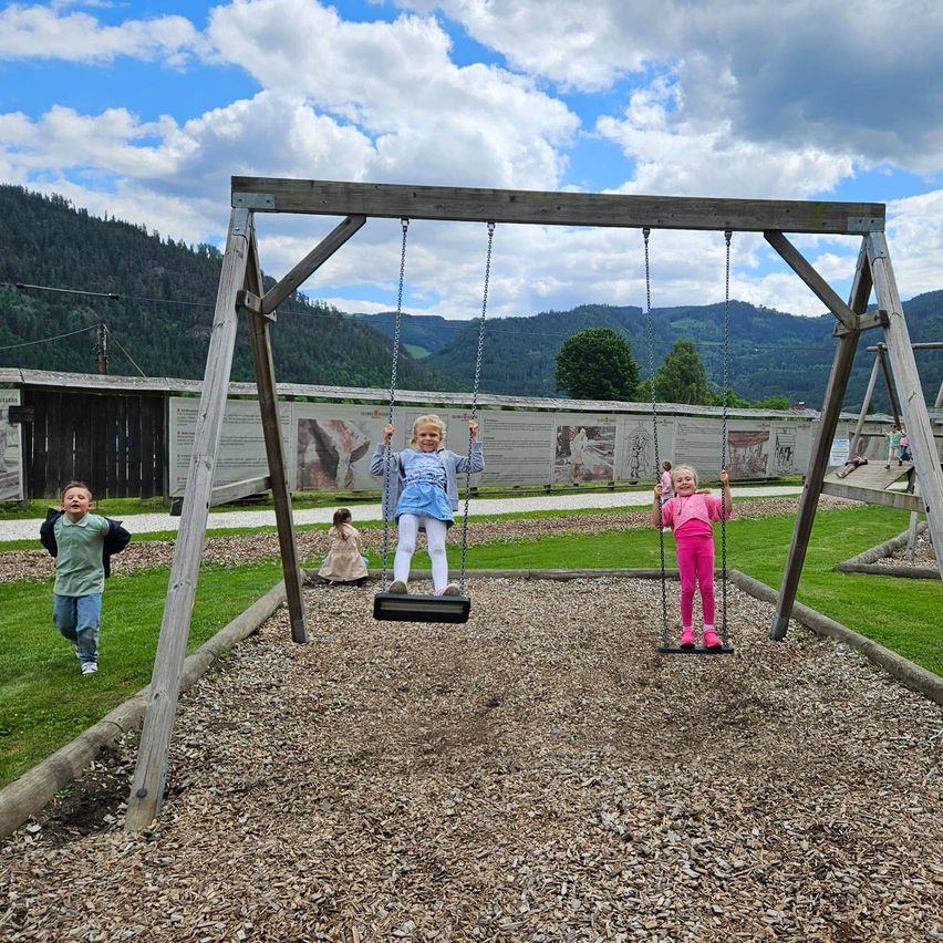 Drei Kinder spielen auf Schaukeln in einem Spielplatz. Die Schaukeln sind aus Holz. Dahinter befinden sich Berge und ein Zaun.
