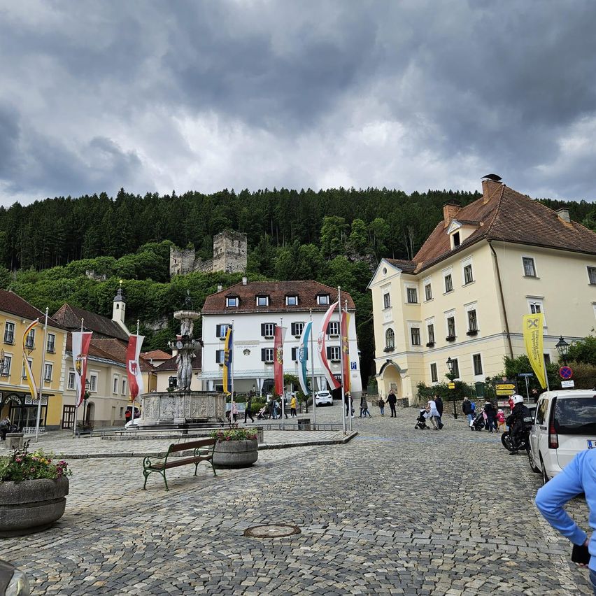 Ein gepflasterter Platz mit einem Brunnen, umgeben von Gebäuden und Fahnen, unter einem bewölkten Himmel, mit einer Burg auf einem Hügel im Hintergrund.