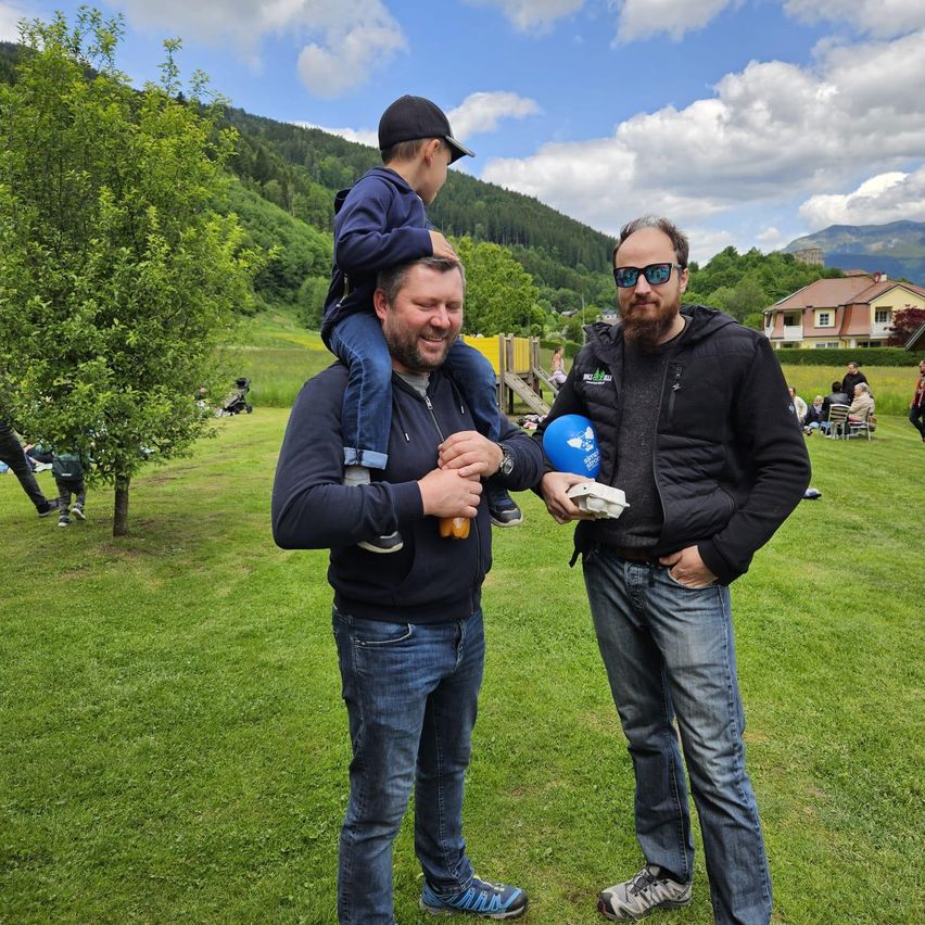 Zwei Männer und ein junger Junge stehen in einem Park. Der Junge sitzt auf den Schultern eines Mannes, während der andere einen blauen Luftballon hält. Sie lächeln und scheinen den Tag zu genießen.