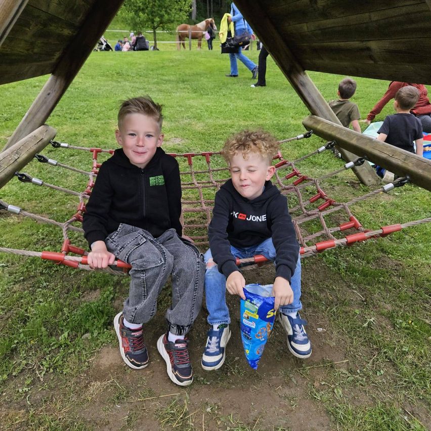 Zwei junge Jungen sitzen auf einer Netzschaukel in einem Park. Ein Junge hält eine Tüte mit Snacks. Hinter ihnen ist ein Grasfeld mit Menschen und Pferden zu sehen.