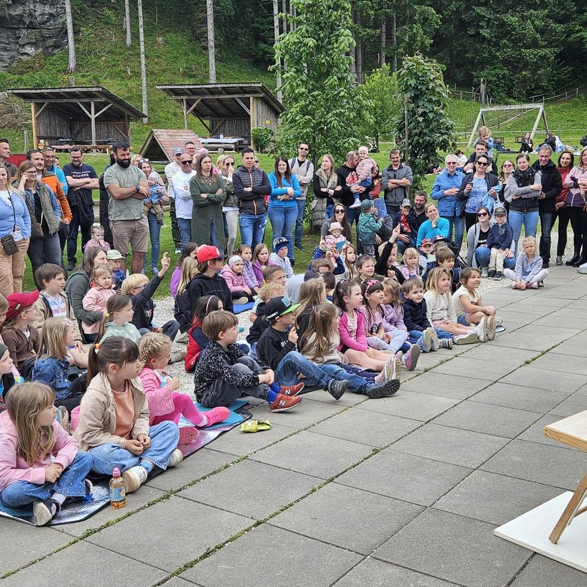 Eine große Gruppe von Menschen, darunter viele Kinder, sitzen im Kreis. Einige stehen hinter ihnen, und einige halten Babys. Rechts befindet sich ein Holztisch.