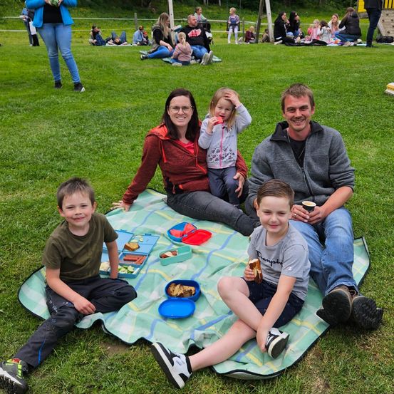 Eine Familie mit fünf Personen, darunter zwei Erwachsene und drei Kinder, sitzt auf einer Decke in einem Park. Sie genießen ein Picknick mit verschiedenen Speisen und Getränken. Die Familie lächelt und scheint eine gute Zeit zu haben.