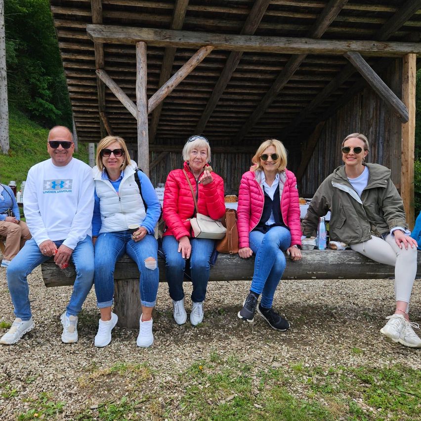 Fünf Erwachsene sitzen auf einer Holzbank vor einer Holzstruktur. Sie tragen Sonnenbrillen und Freizeitkleidung. Bäume und Gras umgeben das Gebiet.