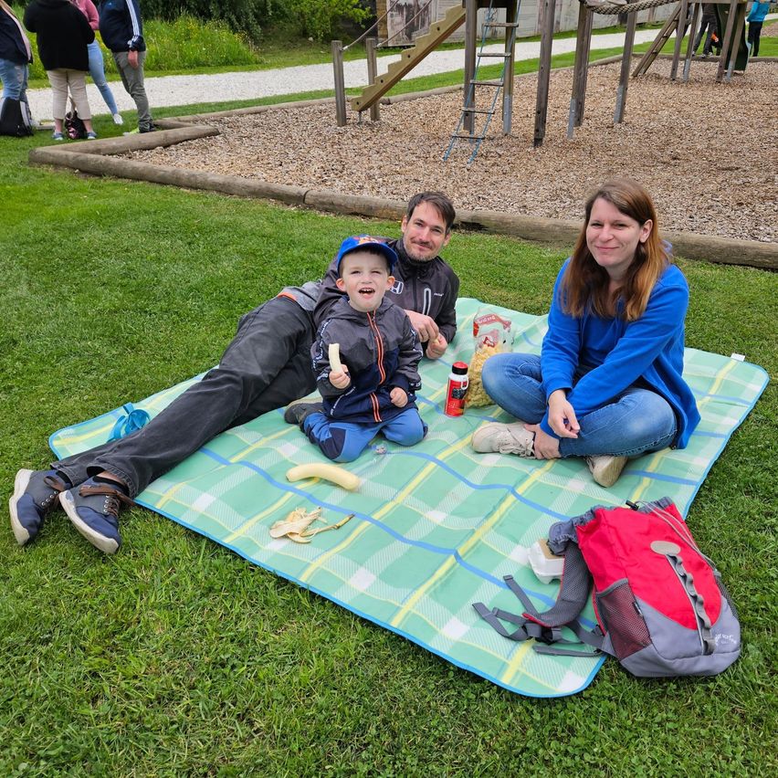 Eine Familie genießt ein Picknick auf einem Rasenfeld mit einem Spielplatz im Hintergrund. Der Mann, die Frau und das Kind sitzen auf einer Decke, lächeln und essen.