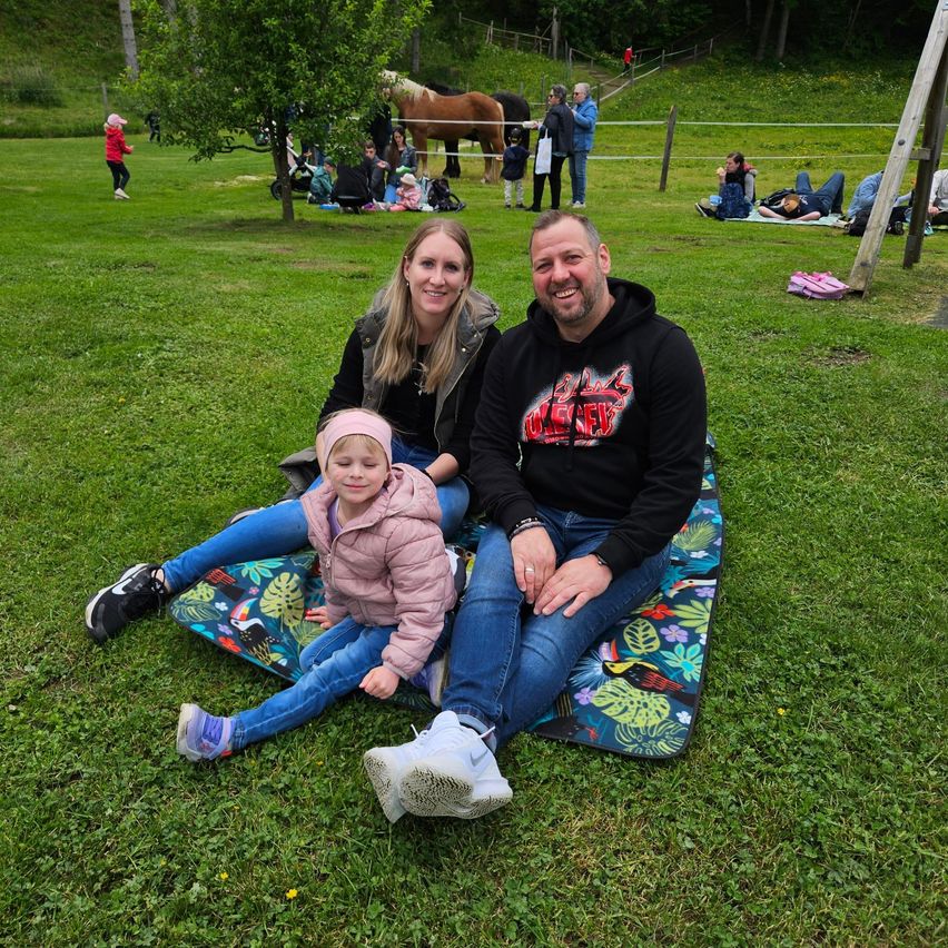 Eine dreiköpfige Familie sitzt auf einer Decke im Park. Das Mädchen trägt eine rosa Jacke, und die Erwachsenen lächeln. Hinter ihnen sitzen und stehen Menschen auf dem Rasen. Ein Pferd ist an einen Zaun gebunden.