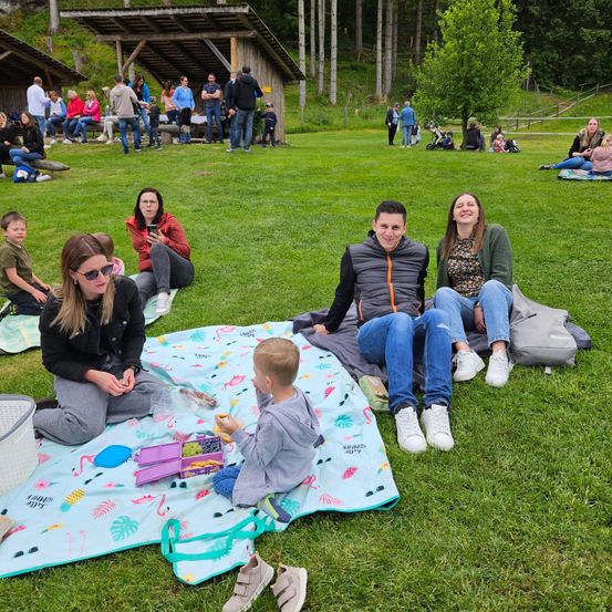 Eine Gruppe von Menschen macht ein Picknick auf einem Rasenfeld. Einige sitzen auf Decken, während andere herumstehen. Eine Frau hält ein Kind, und ein Mann lächelt.