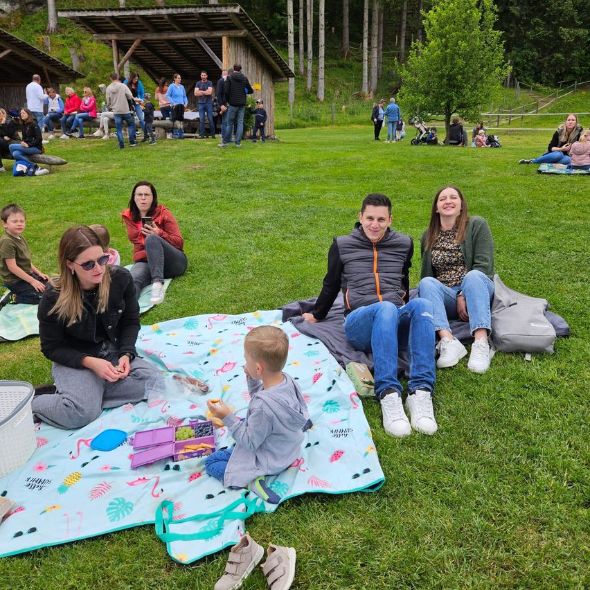 Eine Gruppe von Menschen macht ein Picknick auf einem Rasenfeld. Einige sitzen auf Decken, während andere herumstehen. Eine Frau hält ein Kind, und ein Mann lächelt.