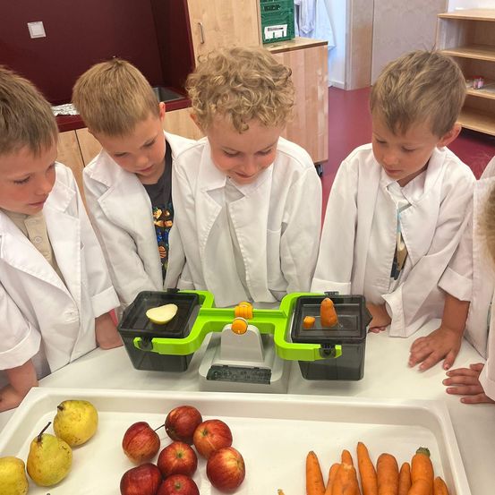Fünf Jungen in weißen Laborkittel lehnen sich über einen Tisch mit Obst und Gemüse. Sie verwenden einen Obstschneider, um Äpfel und Karotten zu schneiden.