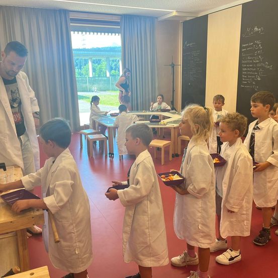 Mehrere junge Kinder in weißen Laborkittel stehen in einem Klassenzimmer mit einem Lehrer. Sie halten Tabletts mit Essen. Dahinter steht eine Tafel mit Schrift und ein Tisch mit sitzenden Kindern.