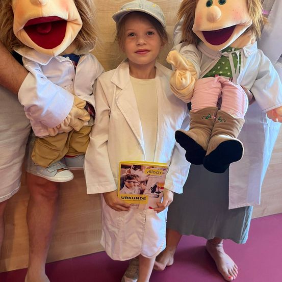 Ein junges Mädchen in einer weißen Laborkittel steht zwischen zwei Puppen und hält ein Buch. Die Puppen sind in weißen Laborkitteln gekleidet und scheinen an einer verspielten Interaktion beteiligt zu sein.