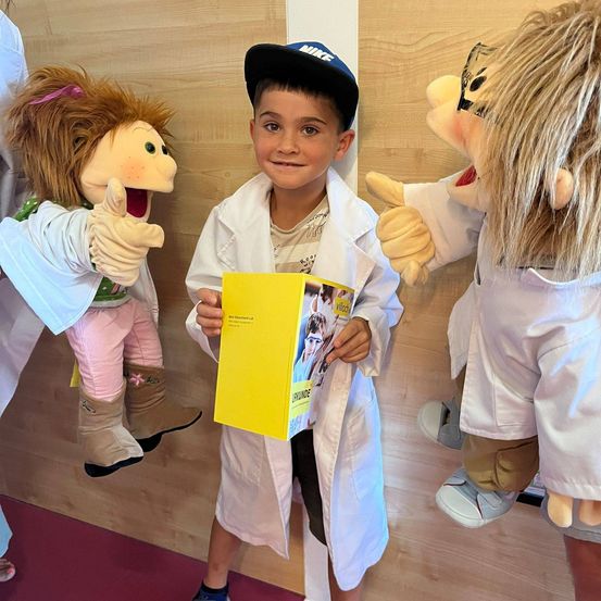 Ein junger Junge in einem weißen Laborkittel hält ein gelbes Buch in der Hand, während er zwischen zwei Puppen steht, die ebenfalls weiße Kittel tragen. Die Puppen haben lange blonde Haare und halten ihre Daumen hoch.