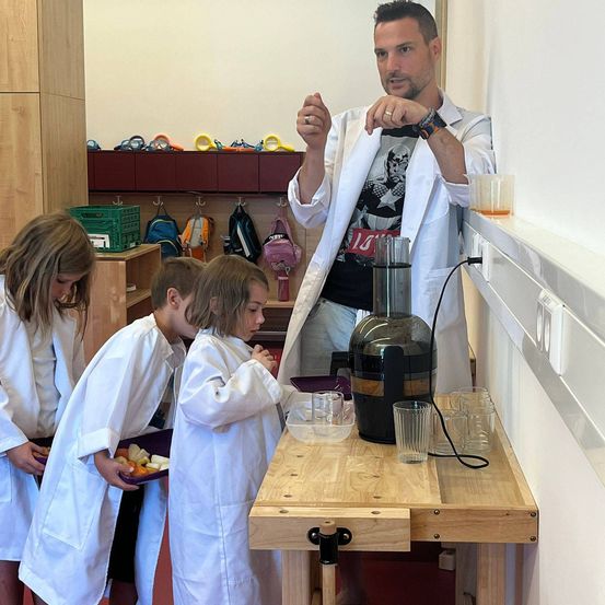 Ein Mann in einem Laborkittel demonstriert drei Kindern in weißen Laborkitteln etwas. Sie schauen auf einen Tisch mit einem Entsafter, Gläsern und Essen.