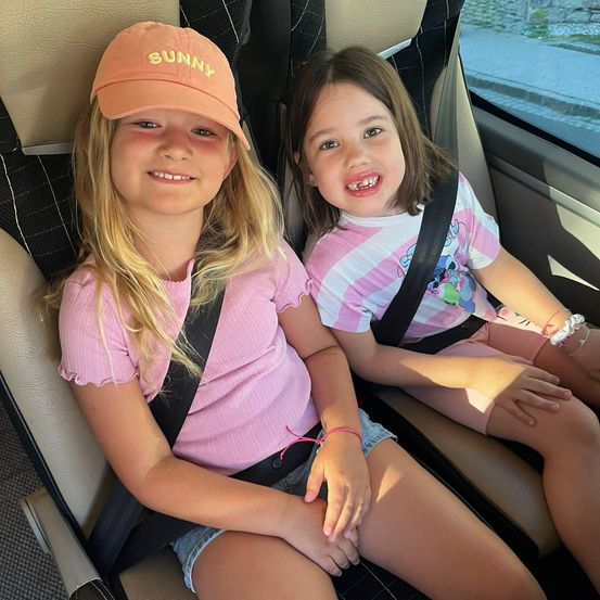 Zwei junge Mädchen sitzen im Rücksitz eines Autos und lächeln. Eine trägt ein rosa Shirt und eine 'Sunny'-Kappe, die andere ein rosa-weiß gestreiftes Shirt. Beide tragen Sicherheitsgurte.