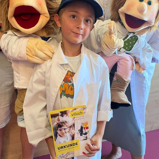 Ein junger Junge in einem weißen Laborkittel steht mit Puppen und hält ein gelbes Buch mit dem Titel 'Urkunde'. Die Puppen tragen Laborkittel, eine mit einem roten Mund, und sie sind alle in einem Raum mit Holzböden.
