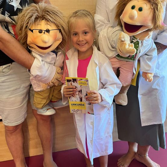 Ein junges Mädchen in einem weißen Laborkittel hält ein Buch und steht zwischen zwei Puppen, die ebenfalls weiße Kittel tragen. Die Puppen halten ein Buch und lächeln. Dahinter steht eine Person in Shorts.