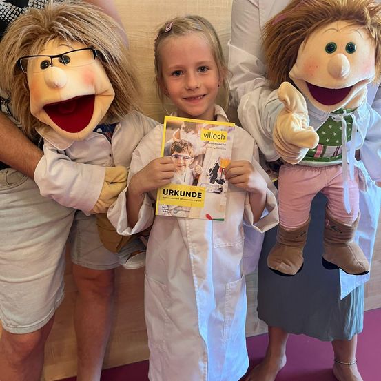 Ein junges Mädchen in einer weißen Laborkittel hält ein Buch, während es zwischen zwei Puppen steht. Eine Puppe trägt eine Brille und die andere hat rosa Hosen und braune Stiefel.