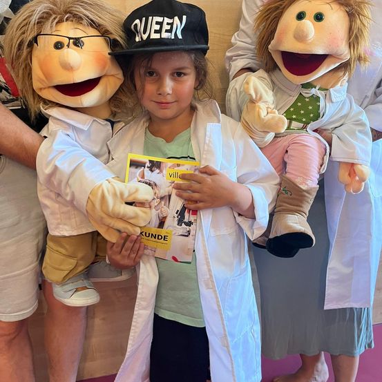 Ein junges Mädchen in einem weißen Laborkittel hält eine Zeitschrift und wird von Puppen umgeben, darunter eine mit einer schwarzen Kappe, die 'QUEEN' beschriftet ist.