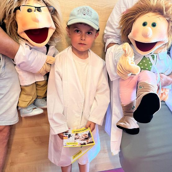 Ein junger Junge in einem weißen Laborkittel steht zwischen zwei Puppen, die ebenfalls weiße Kittel tragen, und hält ein Buch. Die Puppen sind animiert und lächeln.