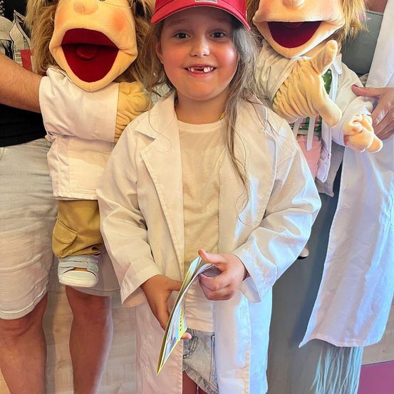 Ein junges Mädchen in einem weißen Laborkittel und einer roten Kappe posiert mit zwei Puppen in Laborkitteln hinter ihr.