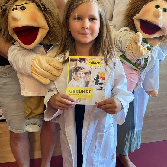 Ein junges Mädchen in einer weißen Laborkittel hält eine Zeitschrift mit dem Titel 'Urkunde' mit einem Puppen hinter sich. Die Puppe trägt eine Brille und hält ein Buch. Eine weitere Puppe steht neben ihr.
