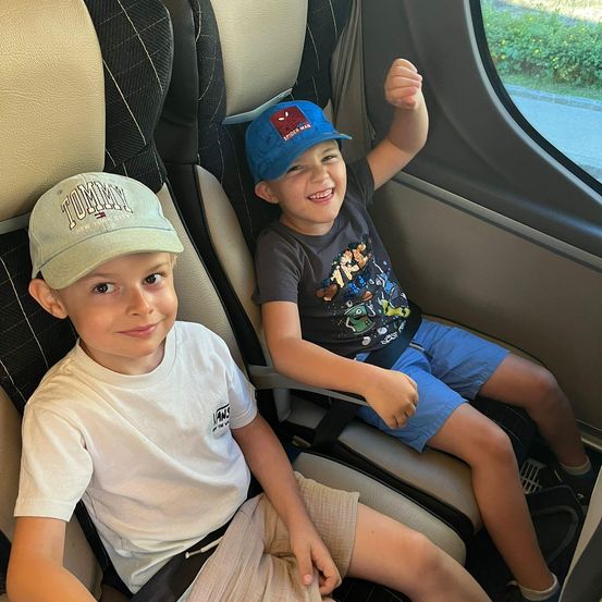 Zwei Jungen sitzen in einem Fahrzeug, einer trägt ein weißes T-Shirt und Khaki-Hosen, der andere eine Spider-Man-Kappe und blaue Shorts. Sie lächeln und scheinen die Fahrt zu genießen.