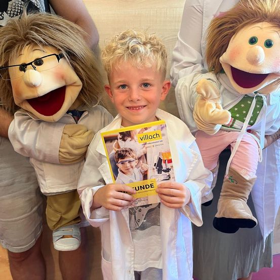 Ein Junge in einem Laborkittel hält ein Buch mit dem Titel 'Unde' und steht zwischen zwei Puppen, die ebenfalls Laborkittel tragen.