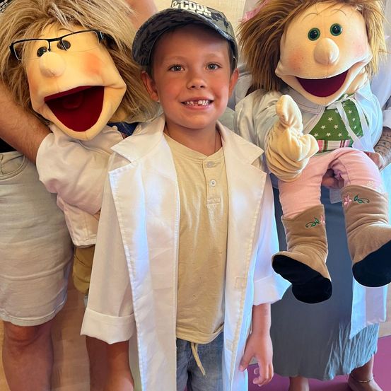 Ein junger Junge steht zwischen zwei Puppen und lächelt für ein Foto. Die Puppen tragen eine Brille und halten eine Babypuppe. Der Junge trägt einen weißen Laborkittel und eine Kappe.