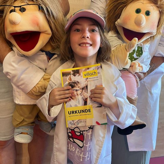 Ein junges Mädchen in einem Laborkittel hält ein Buch in der Hand, lächelt und wird von zwei Marionetten in Laborkitteln umgeben, von denen eine eine Brille trägt und die andere grüne Augen hat.