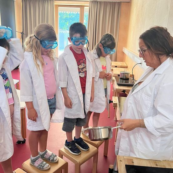 Eine Frau in einer Laborkittel hält einen Topf, während Kinder in Laborkitteln zusehen. Sie stehen auf Hockern in einem Klassenzimmer mit einem Herd und einem Fenster.