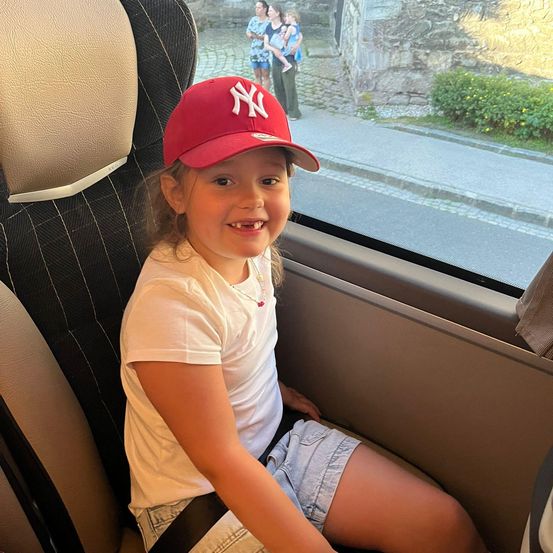 Ein junges Mädchen mit einer NY-Baseballkappe lächelt in einem Fahrzeug, mit einer gepflasterten Straße und Menschen im Hintergrund.