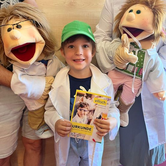 Ein Kind hält ein Buch über ein Bildungslabor in der Hand, während es mit zwei Puppen in weißen Laborkittel posiert. Eine Puppe trägt eine Brille und die andere einen grünen Hut.