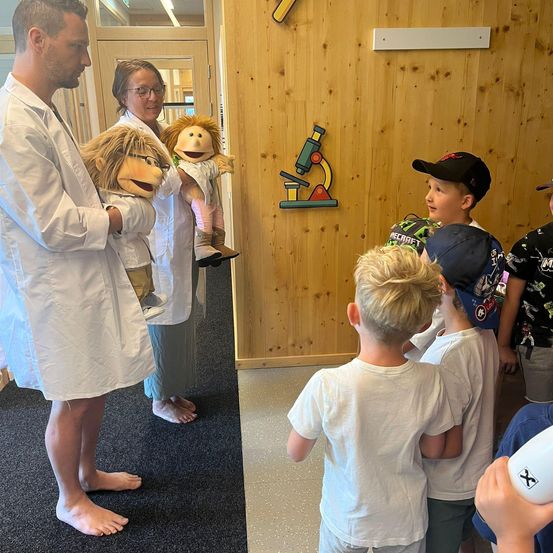 Zwei Personen in Laborkittel halten Puppen in einem Raum mit Holzwänden. Kinder beobachten, einer hält eine Flasche, während eine Person im Hintergrund eine Brille trägt.
