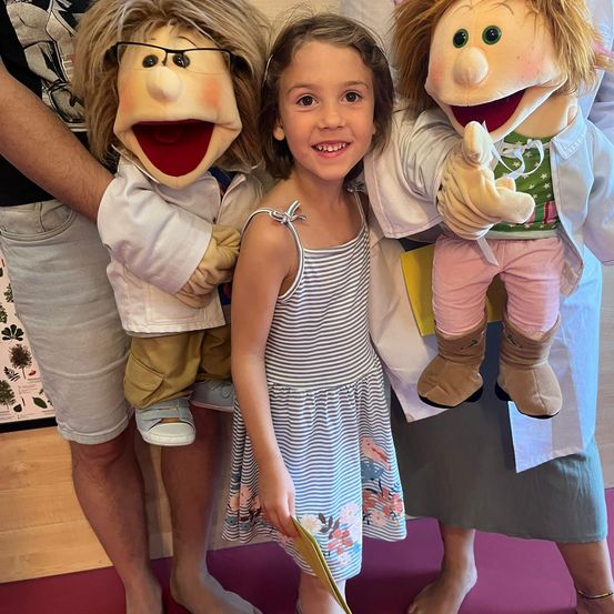Ein Mädchen in einem gestreiften Kleid posiert mit zwei Puppen, eine mit Brille und Laborkittel, die andere in einem grünen Shirt und rosa Hosen. Die Puppen werden von Erwachsenen gehalten.