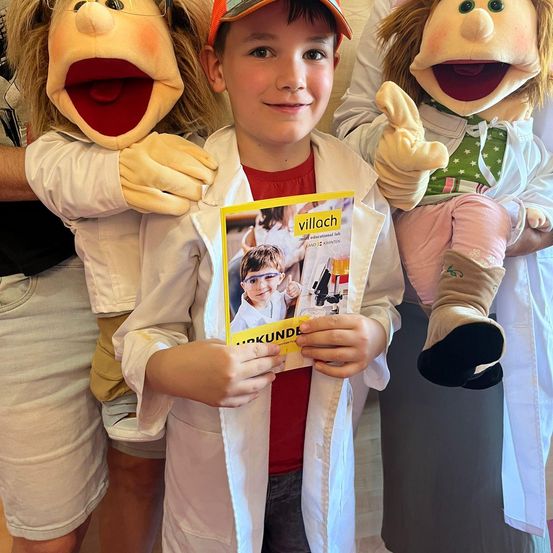 Ein junger Junge in einem Laborkittel hält ein Buch, während zwei Puppen in weißen Kitteln hinter ihm stehen. Das Buch zeigt ein Bild eines Kindes mit Brille und Laborkittel.