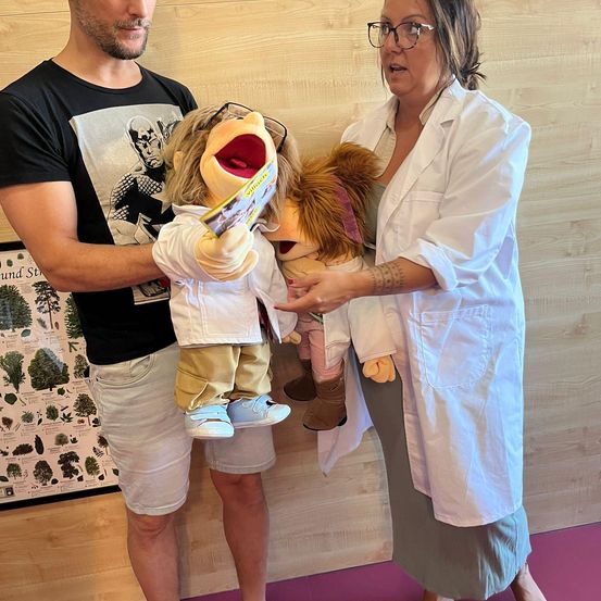 Ein Mann und eine Frau in weißen Kitteln halten Puppen in einem Raum. Die Puppen tragen eine Brille und halten ein Buch. Die Frau spricht. Hinter ihnen ist eine Holzwand mit einem Baumdiagramm.